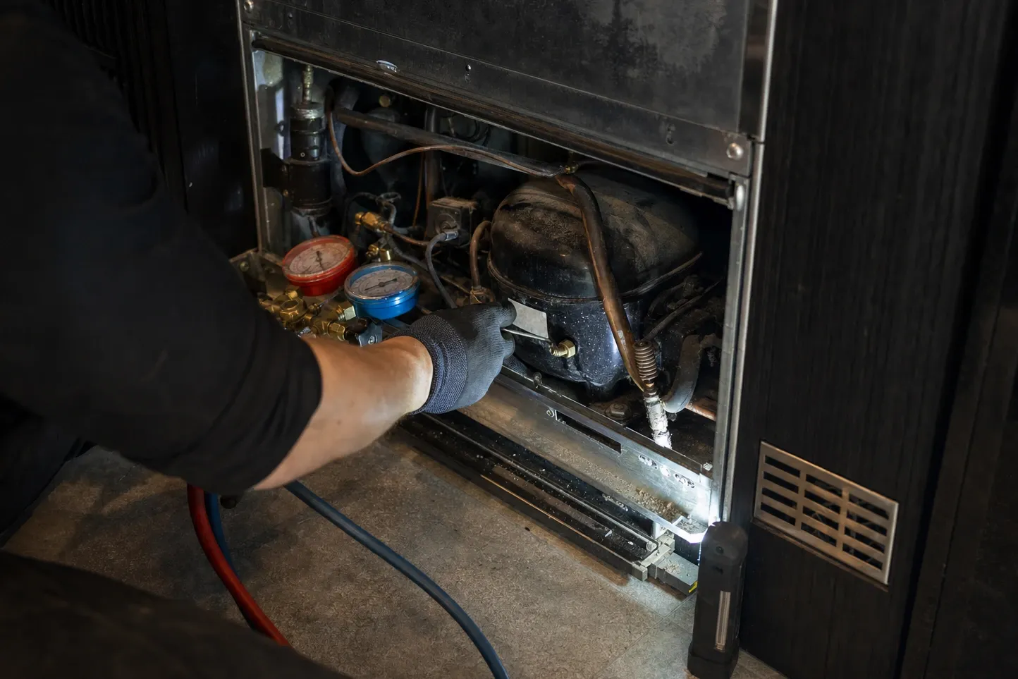 Gloved hand holding manifold gauges at the compressor behind the lower grille, work light on the floor
